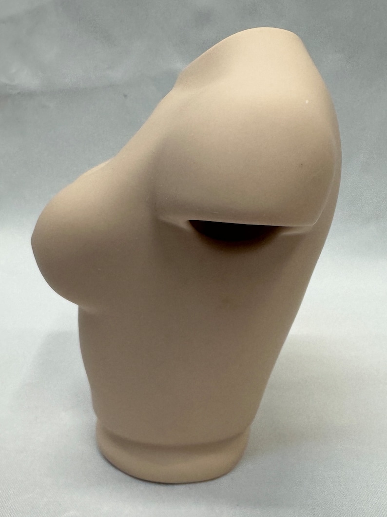 May include: A beige ceramic bust sculpture with a smooth, rounded surface. The bust is missing the head and neck, and the shoulders are sculpted in a relaxed pose.