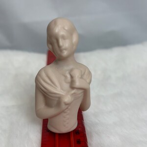Może przedstawiać: Mała, kremowa figurka ceramiczna przedstawiająca kobietę w szalu trzymającą kwiat. Figurka ma około 10 cm wysokości i stoi na czerwonej linijce.