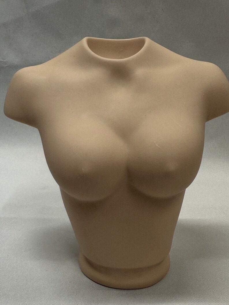 May include: A beige ceramic bust of a person's torso. The bust is smooth and has a rounded shape.