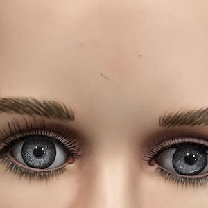 Pode incluir: Close-up do rosto de um manequim, com foco nos olhos. Os olhos são de uma cor cinza marcante com cílios longos e escuros e sobrancelhas bem definidas. O tom de pele é pálido e neutro.