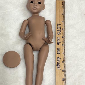 Puede incluir: Una muñeca de piel bronceada con extremidades articuladas y cabeza calva, tumbada sobre una superficie blanca. Una regla de madera con el texto "LETS rule out drugs!" está al lado de la muñeca. También hay una base redonda de color marrón.