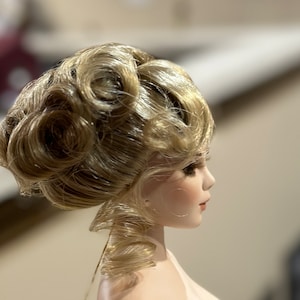 Op de afbeelding: Een porseleinen pop met een blond pruikje dat is gestyled in een hoge knot met krullen. De pop heeft een geschilderd gezicht met bruine ogen en roze lippen.