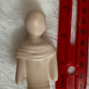 Może przedstawiać: Mała, beżowa figurka ceramiczna przedstawiająca osobę w koszuli z długim rękawem. Figurka ma około 10 cm wysokości i stoi na białym tle.