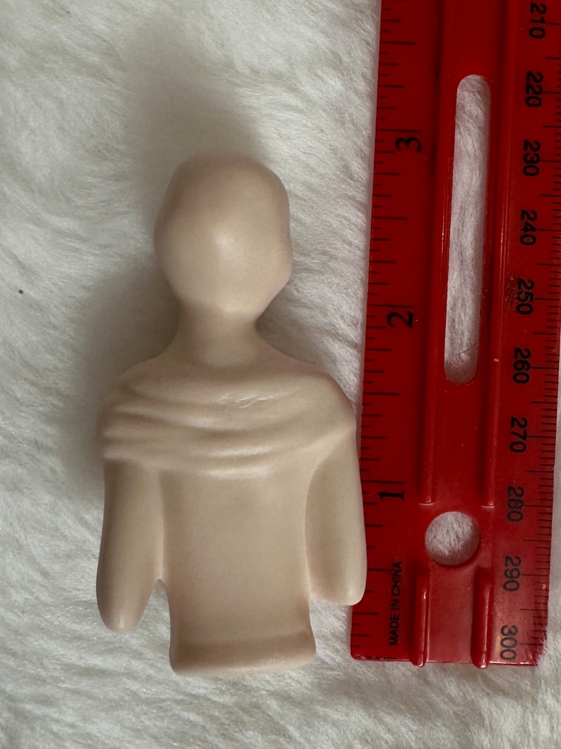 Może przedstawiać: Mała, beżowa figurka ceramiczna przedstawiająca osobę w koszuli z długim rękawem. Figurka ma około 10 cm wysokości i stoi na białym tle.