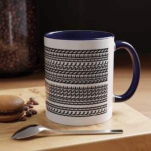 May include: White ceramic mug with a navy blue interior and handle. The mug features a repeating black geometric pattern. A spoon, a macaron, and coffee beans are on a wooden surface next to the mug.