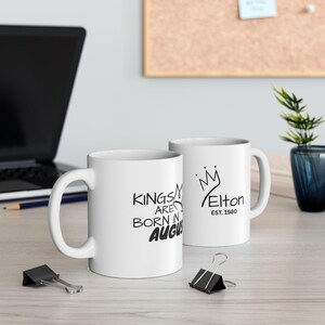 May include: Two white ceramic mugs. One mug has the text "KINGS ARE BORN IN AUGUST" in black. The other mug has a crown design with the name "Elton" and "EST. 1980" in black. Both mugs have a curved handle.