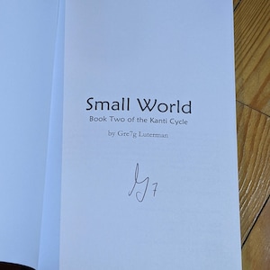 Op de afbeelding: Een wit boek met de titel "Small World" en de ondertitel "Book Two of the Kanti Cycle" door Greg Lutherman. Het boek is gesigneerd met een handgeschreven handtekening.