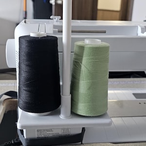 May include: A sewing machine with a white thread stand holding a black and a pale green spool of thread. The machine is on a light grey surface.