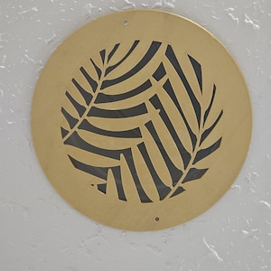 Puede incluir: Una rejilla de ventilación redonda de metal dorado con un diseño de hoja negra.