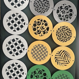 Op de afbeelding: Een verzameling van 16 ronde afvoerdeksels met verschillende geometrische en bloemmotieven. De deksels zijn gemaakt van wit, grijs, goud en groen materiaal.