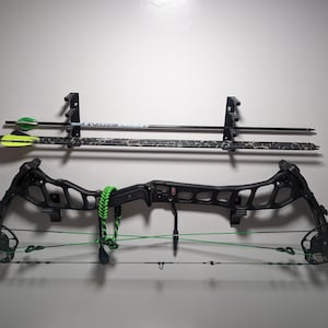 Peut inclure: Un arc à poulies noir avec une corde verte et un carquois de flèches noir et vert est monté sur un mur blanc. Deux flèches noires et blanches sont également montées sur le mur.