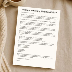 May include: A devotional titled "Welcome to Raising Kingdom Kids&trade;" with text on a white paper. A cup of tea sits to the left. The text is in black font. The devotional is on a cream-colored surface.