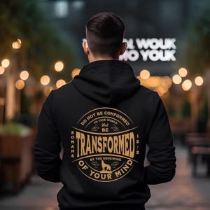 Pode incluir: Camisola de capuz preta com um gráfico circular dourado nas costas. O gráfico apresenta o texto "Não se conformem a este mundo, mas deixem-se transformar pela renovação da sua mente" com a referência bíblica "Romanos 12:2".