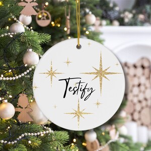 May include: White round Christmas ornament with gold glitter stars and the word "Testify" written in black cursive.