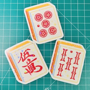 May include: Three rectangular stickers featuring Mahjong tiles. The tiles have a white background with red symbols and designs. The edges are outlined in orange and gold. The stickers have a holographic, glittery effect.