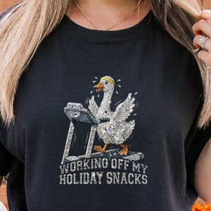 May include: Black t-shirt featuring a goose on a treadmill with the text "WORKING OFF MY HOLIDAY SNACKS". The goose is wearing a yellow hat. The shirt is a dark color and has a relaxed fit.