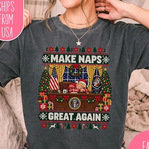 May include: A charcoal gray short-sleeve t-shirt featuring a graphic print of a person in a red sweater and Santa hat sleeping at a desk. The text reads MAKE NAPS GREAT AGAIN. The design includes Christmas trees, flags, and holiday patterns.