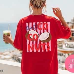 May include: A red t-shirt with the text "that's CO NUTS" and a coconut graphic. The design is on the back of the shirt, with pink and white vertical stripes. The shirt is a casual, oversized style.