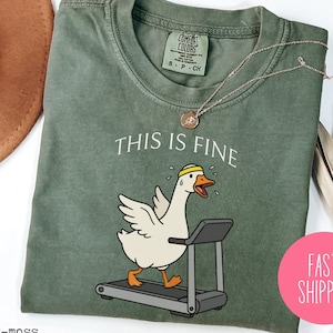 May include: A sage green t-shirt with the phrase "THIS IS FINE" above a cartoon goose running on a treadmill. The goose is wearing a yellow headband. A gold necklace and a pink "FAST SHIPPING" sticker are also visible.