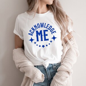 May include: White t-shirt with a blue circle graphic that says "Acknowledge Me" with stars around the circle.