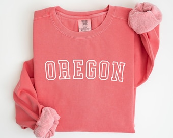 May include: A coral pink sweatshirt with the word "OREGON" in white outlined letters. The sweatshirt is made by "COMFORT COLORS" and has a crew neck.