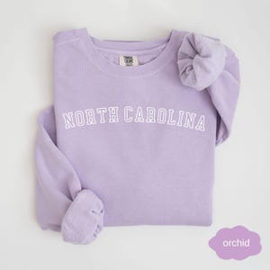 May include: A lavender sweatshirt with the text "NORTH CAROLINA" in white on the front. The sweatshirt is made of a soft, comfortable material and has a relaxed fit.