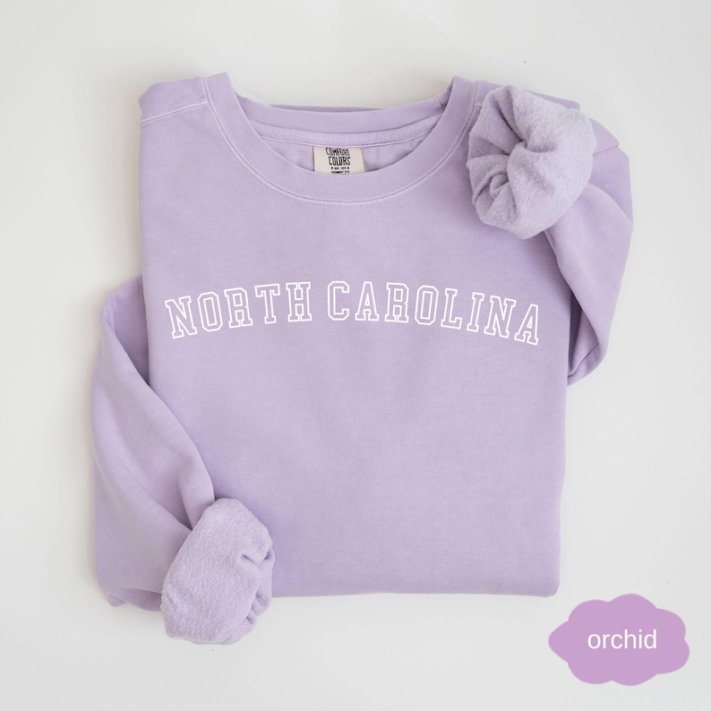May include: A lavender sweatshirt with the text "NORTH CAROLINA" in white on the front. The sweatshirt is made of a soft, comfortable material and has a relaxed fit.