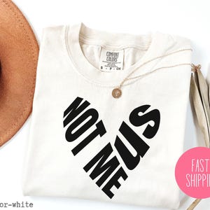 May include: White t-shirt with black text that reads "Not Me Us" in a heart shape.