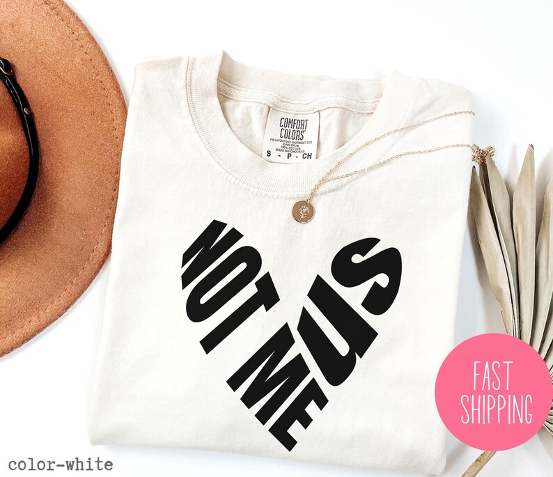 May include: White t-shirt with black text that reads "Not Me Us" in a heart shape.
