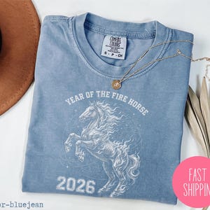 K&ouml;nnte beinhalten: Hellblaues T-Shirt mit einem wei&szlig;en Motiv eines Pferdes und dem Text "YEAR OF THE FIRE HORSE 2026". Das T-Shirt hat einen Rundhalsausschnitt und kurze &Auml;rmel. Eine goldene Halskette liegt auf dem T-Shirt. Ein rosa Kreis mit "FAST SHIPPING" ist im Bild.