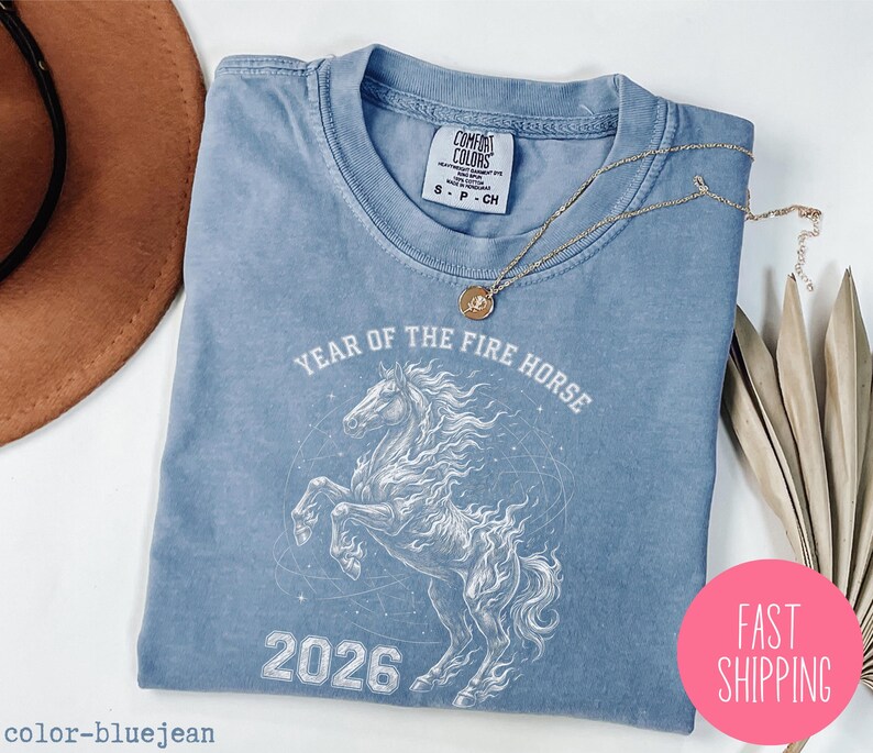 K&ouml;nnte beinhalten: Hellblaues T-Shirt mit einem wei&szlig;en Motiv eines Pferdes und dem Text "YEAR OF THE FIRE HORSE 2026". Das T-Shirt hat einen Rundhalsausschnitt und kurze &Auml;rmel. Eine goldene Halskette liegt auf dem T-Shirt. Ein rosa Kreis mit "FAST SHIPPING" ist im Bild.