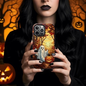 May include: A Halloween-themed phone case featuring a stained-glass design with a ghost and jack-o'-lantern. The case has a brown and orange color scheme with a forest and sunset background. The phone case is held in front of a blurred orange background.