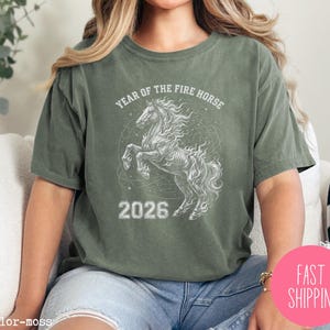 K&ouml;nnte beinhalten: Olivgr&uuml;nes T-Shirt mit wei&szlig;er Grafik eines steigenden Pferdes und dem Text "YEAR OF THE FIRE HORSE 2026". Das Shirt hat eine lockere Passform.