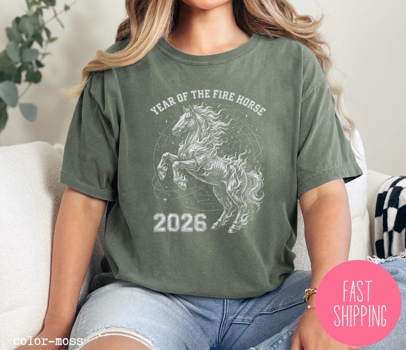 K&ouml;nnte beinhalten: Olivgr&uuml;nes T-Shirt mit wei&szlig;er Grafik eines steigenden Pferdes und dem Text "YEAR OF THE FIRE HORSE 2026". Das Shirt hat eine lockere Passform.