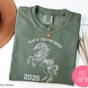 K&ouml;nnte beinhalten: Ein salbeigr&uuml;nes T-Shirt mit einer Grafik eines sich aufb&auml;umenden Pferdes, dem Text "YEAR OF THE FIRE HORSE 2026" und einem Comfort Colors-Etikett. Eine goldene Halskette und ein pinkfarbener "FAST SHIPPING"-Aufkleber sind ebenfalls sichtbar.