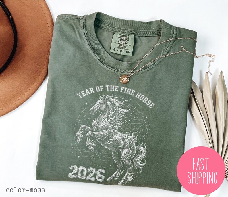 K&ouml;nnte beinhalten: Ein salbeigr&uuml;nes T-Shirt mit einer Grafik eines sich aufb&auml;umenden Pferdes, dem Text "YEAR OF THE FIRE HORSE 2026" und einem Comfort Colors-Etikett. Eine goldene Halskette und ein pinkfarbener "FAST SHIPPING"-Aufkleber sind ebenfalls sichtbar.