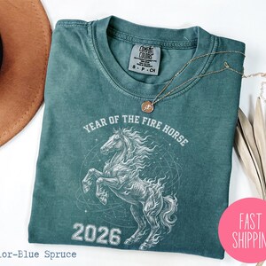 K&ouml;nnte beinhalten: Blaugr&uuml;nes T-Shirt mit einem wei&szlig;en Pferdemotiv, dem Text "YEAR OF THE FIRE HORSE 2026" und einem Comfort Colors-Etikett. Eine goldene Halskette und ein pinkfarbener "FAST SHIPPING"-Aufkleber sind ebenfalls zu sehen.