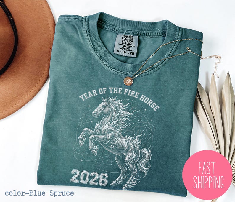 K&ouml;nnte beinhalten: Blaugr&uuml;nes T-Shirt mit einem wei&szlig;en Pferdemotiv, dem Text "YEAR OF THE FIRE HORSE 2026" und einem Comfort Colors-Etikett. Eine goldene Halskette und ein pinkfarbener "FAST SHIPPING"-Aufkleber sind ebenfalls zu sehen.