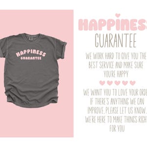 K&ouml;nnte beinhalten: Ein graues T-Shirt mit dem Text "HAPPINESS GUARANTEE" in rosa Buchstaben. Das Shirt wird auf einem rosa Hintergrund angezeigt. Der Text auf der rechten Seite des Bildes lautet "HAPPINESS GUARANTEE".
