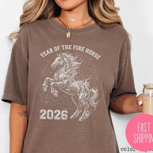K&ouml;nnte beinhalten: Braunes T-Shirt mit einer wei&szlig;en Illustration eines sich aufb&auml;umenden Pferdes mit feuriger M&auml;hne und dem Text "YEAR OF THE FIRE HORSE 2026". Das Design ist auf dem T-Shirt zentriert.