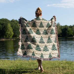 May include: A person holding a woven throw blanket with a green and white Christmas tree pattern. The blanket has fringe on the edges.