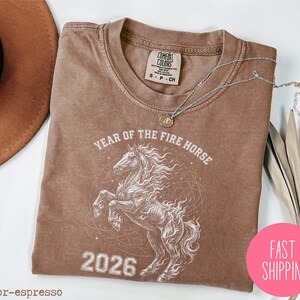 K&ouml;nnte beinhalten: Espressofarbenes T-Shirt mit wei&szlig;er Grafik eines steigenden Pferdes, dem Text "YEAR OF THE FIRE HORSE 2026" und einem pinkfarbenen "FAST SHIPPING" Aufkleber. Eine silberne Halskette mit Anh&auml;nger liegt auf dem Shirt.