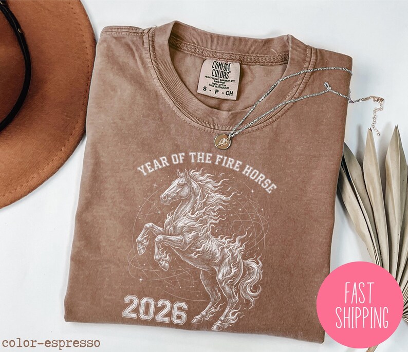 K&ouml;nnte beinhalten: Espressofarbenes T-Shirt mit wei&szlig;er Grafik eines steigenden Pferdes, dem Text "YEAR OF THE FIRE HORSE 2026" und einem pinkfarbenen "FAST SHIPPING" Aufkleber. Eine silberne Halskette mit Anh&auml;nger liegt auf dem Shirt.