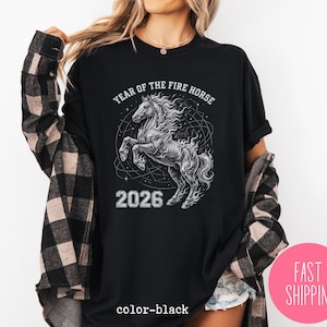 K&ouml;nnte beinhalten: Schwarzes T-Shirt mit einer detaillierten Grafik eines sich aufb&auml;umenden Pferdes, umgeben von einem Himmelsdesign. Der Text "YEAR OF THE FIRE HORSE" und "2026" sind oberhalb und unterhalb des Pferdes aufgedruckt. Die Worte "color-black" sind unten aufgedruckt.