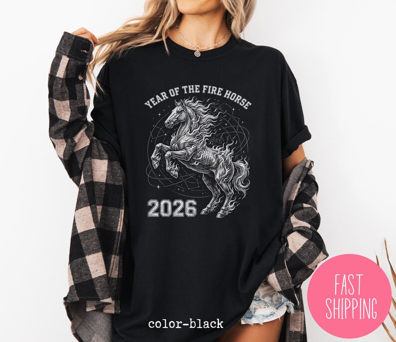 K&ouml;nnte beinhalten: Schwarzes T-Shirt mit einer detaillierten Grafik eines sich aufb&auml;umenden Pferdes, umgeben von einem Himmelsdesign. Der Text "YEAR OF THE FIRE HORSE" und "2026" sind oberhalb und unterhalb des Pferdes aufgedruckt. Die Worte "color-black" sind unten aufgedruckt.