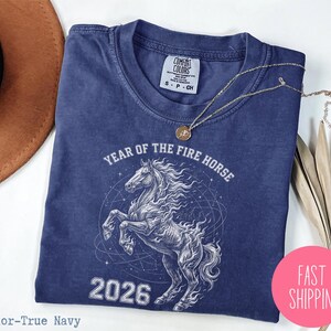 K&ouml;nnte beinhalten: Marineblaues T-Shirt mit wei&szlig;er Grafik eines steigenden Pferdes, dem Text "YEAR OF THE FIRE HORSE" und der Zahl "2026". Das Shirt hat einen Rundhalsausschnitt und kurze &Auml;rmel. Eine goldene Halskette liegt auf dem Shirt.