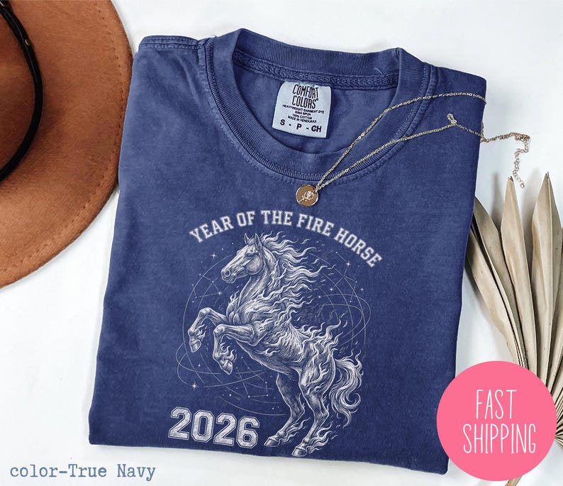 K&ouml;nnte beinhalten: Marineblaues T-Shirt mit wei&szlig;er Grafik eines steigenden Pferdes, dem Text "YEAR OF THE FIRE HORSE" und der Zahl "2026". Das Shirt hat einen Rundhalsausschnitt und kurze &Auml;rmel. Eine goldene Halskette liegt auf dem Shirt.