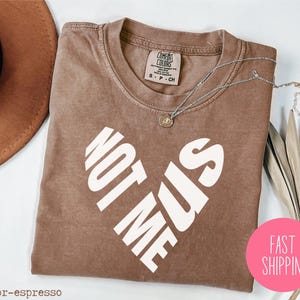 May include: A brown t-shirt with the text "Not Me Sis" printed in white in a heart shape.