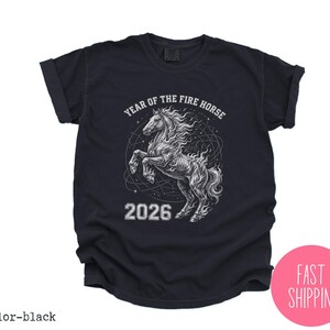 K&ouml;nnte beinhalten: Schwarzes T-Shirt mit einer Grafik eines sich aufb&auml;umenden Pferdes mit flie&szlig;ender M&auml;hne, umgeben von einem himmlischen Design. Der Text "YEAR OF THE FIRE HORSE" und "2026" ist &uuml;ber und unter der Pferdeillustration gedruckt.