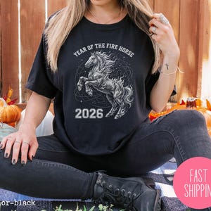 K&ouml;nnte beinhalten: Schwarzes T-Shirt mit einer detaillierten Illustration eines sich aufb&auml;umenden Pferdes, der Text lautet "YEAR OF THE FIRE HORSE" und "2026". Das Design ist in Wei&szlig;- und Graut&ouml;nen gehalten. Das T-Shirt ist schwarz.